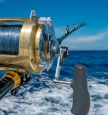 A close-up view of a fishing rod and reel on a boat, angling out over a deep blue ocean with a clear sky and a few clouds. The reel is shiny, metallic, and well-crafted, indicating it is likely for deep-sea fishing.