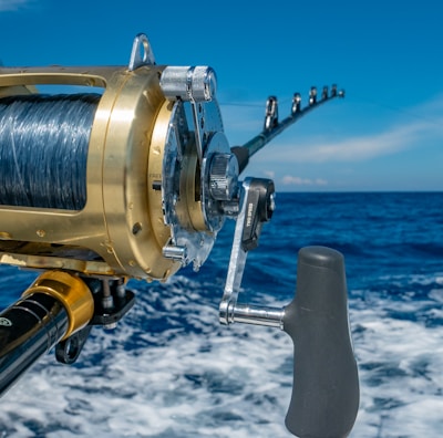 A close-up view of a fishing rod and reel on a boat, angling out over a deep blue ocean with a clear sky and a few clouds. The reel is shiny, metallic, and well-crafted, indicating it is likely for deep-sea fishing.