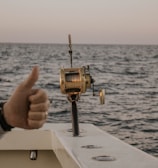 A boat fishing trip on the open sea with a fishing reel mounted on the side of the boat. The ocean stretches to the horizon under a clear sky, and a person gives a thumbs-up gesture, indicating approval or success.