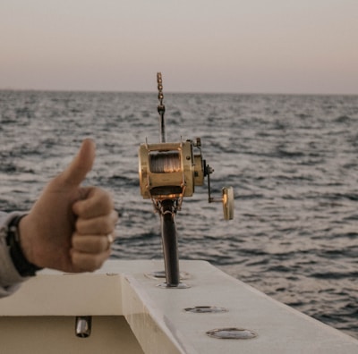 A boat fishing trip on the open sea with a fishing reel mounted on the side of the boat. The ocean stretches to the horizon under a clear sky, and a person gives a thumbs-up gesture, indicating approval or success.