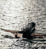 A fishing boat with large nets cast out on the sides floats on the water, with shimmering sunlight reflecting off the rippling surface of the sea.