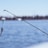 A fishing rod extends into the frame against a backdrop of calm, blue water. The rod is equipped with a line, from which hangs a colorful lure designed to mimic a worm. In the distance, the shoreline is faintly visible, slightly blurred due to the focus on the rod and lure.