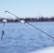 A fishing rod extends into the frame against a backdrop of calm, blue water. The rod is equipped with a line, from which hangs a colorful lure designed to mimic a worm. In the distance, the shoreline is faintly visible, slightly blurred due to the focus on the rod and lure.