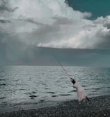 A person in a striped outfit is fishing by the shore, casting a long fishing rod into the sea. The waves gently lap at the pebbled beach, while large, dramatic clouds fill the sky above, creating a contrast with the dark water.