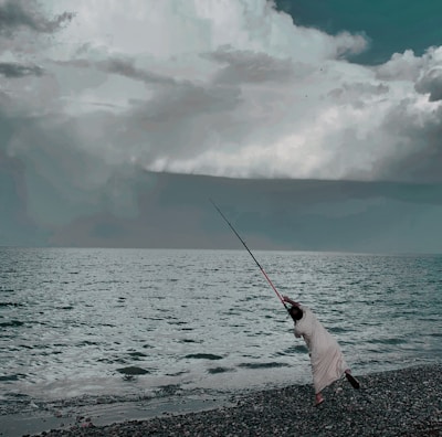 A person in a striped outfit is fishing by the shore, casting a long fishing rod into the sea. The waves gently lap at the pebbled beach, while large, dramatic clouds fill the sky above, creating a contrast with the dark water.