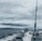 A fishing rod stands upright on a moving boat against a backdrop of cloudy skies and expansive water. The boat motor leaves a trail of white wake behind as it traverses the dark, choppy waters.