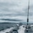 A fishing rod stands upright on a moving boat against a backdrop of cloudy skies and expansive water. The boat motor leaves a trail of white wake behind as it traverses the dark, choppy waters.