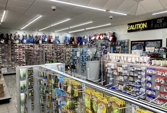 A well-lit store interior filled with fishing and outdoor equipment. The shelves are stocked with various lures, fishing lines, and other accessories. A range of colorful life vests is displayed on the back wall. A caution sign and a digital temperature display are mounted near the ceiling.