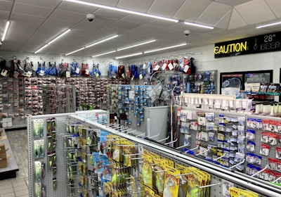 A well-lit store interior filled with fishing and outdoor equipment. The shelves are stocked with various lures, fishing lines, and other accessories. A range of colorful life vests is displayed on the back wall. A caution sign and a digital temperature display are mounted near the ceiling.
