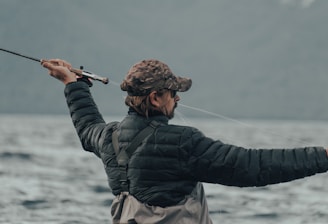 A person wearing a camouflage cap and a black puffy jacket is holding a fishing rod, actively casting a line into a large body of water. The background is composed of blurred mountains and a cloudy sky, conveying an outdoor, nature setting.