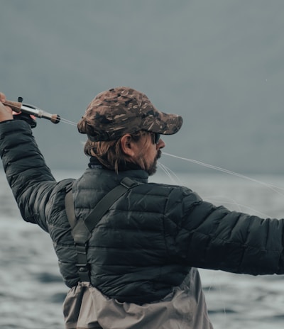 A person wearing a camouflage cap and a black puffy jacket is holding a fishing rod, actively casting a line into a large body of water. The background is composed of blurred mountains and a cloudy sky, conveying an outdoor, nature setting.