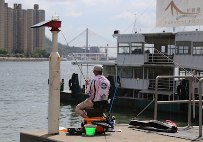 A person is seated on the edge of a pier, fishing beside a large boat marked 'Park Island'. In the background, a prominent bridge and high-rise buildings are visible across the water. Fishing equipment is spread out around the seated individual, who is wearing a cap and patterned shirt.