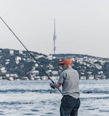 A man wearing a checkered shirt and orange cap is fishing by the waterfront. In the background, a cityscape with numerous buildings along a tree-lined shore is visible. A tall tower rises prominently in the distance against a clear sky.