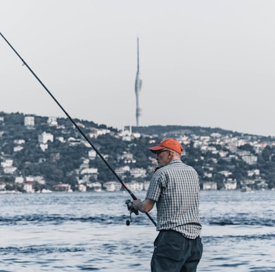 A man wearing a checkered shirt and orange cap is fishing by the waterfront. In the background, a cityscape with numerous buildings along a tree-lined shore is visible. A tall tower rises prominently in the distance against a clear sky.