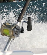 A close-up of a fishing reel mounted on a fishing rod, with a vibrant green line. The background features splashing water, suggesting movement and action, possibly from a boat cutting through the waves.