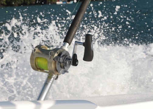 A close-up of a fishing reel mounted on a fishing rod, with a vibrant green line. The background features splashing water, suggesting movement and action, possibly from a boat cutting through the waves.