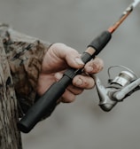 A person wearing a camouflage jacket is holding a fishing rod. The fishing reel is prominently featured, with the hand showing some dirt and wear, suggesting an outdoor activity.