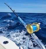 A fishing rod with a gold reel is mounted on the side of a boat, extending over the deep blue ocean. The waves create white foam as the boat moves forward under a clear blue sky.