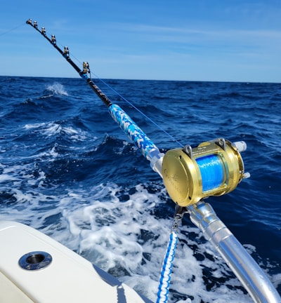 A fishing rod with a gold reel is mounted on the side of a boat, extending over the deep blue ocean. The waves create white foam as the boat moves forward under a clear blue sky.
