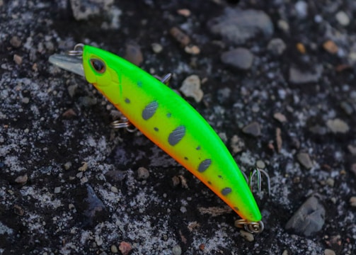 A bright and colorful fishing lure rests on a textured, rocky surface. The lure is predominantly neon green with orange highlights and black spots, equipped with metal hooks.