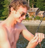 A shirtless man with sunglasses joyfully holds a fishing rod with a small fish caught on the line. In the background, there is a serene lakeside setting with lush green trees and a wooden cabin.