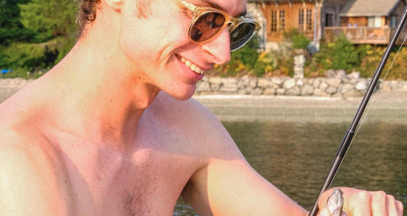 A shirtless man with sunglasses joyfully holds a fishing rod with a small fish caught on the line. In the background, there is a serene lakeside setting with lush green trees and a wooden cabin.
