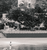 A person is standing on rocks by the edge of a body of water, holding a fishing rod. In the background, there is a sandy area, trees, and several buildings, including a multi-story structure with a tiled roof. The surrounding environment includes vegetation and a few outdoor settings with chairs and tables.